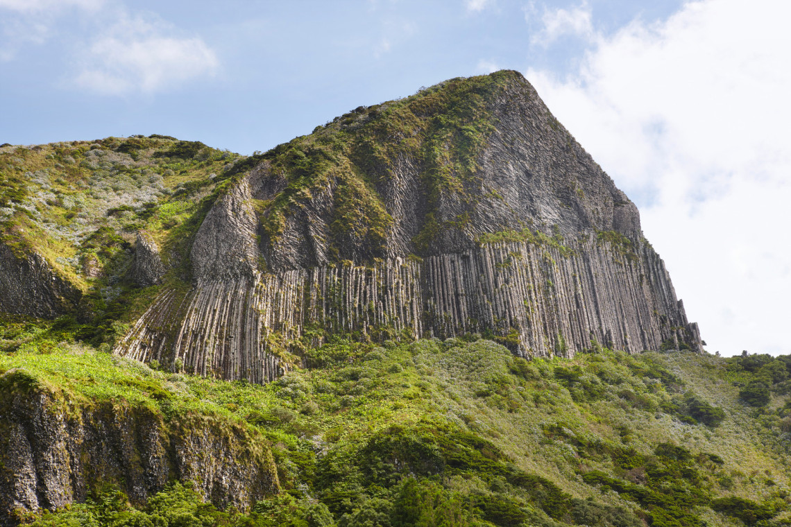 Rocha dos Bord&otilde;es rises above the green landscape of Flores Island in the Azores, Portugal. Its distinctive rock face is formed by tall vertical volcanic columns created through prismatic jointing.