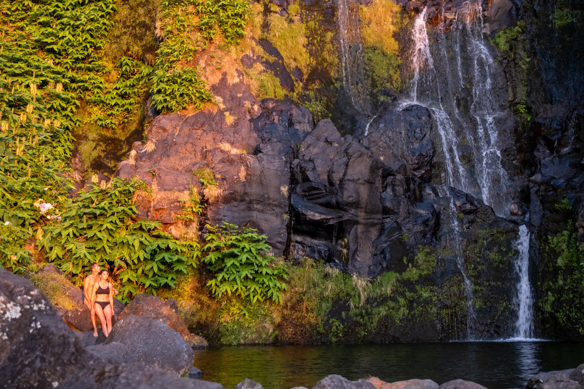 Po&ccedil;o do Bacalhau waterfall cascades over dark volcanic rock into a natural pool in Faj&atilde; Grande on Flores Island. Dense ferns and other vegetation cover the rock walls surrounding the swimming area.