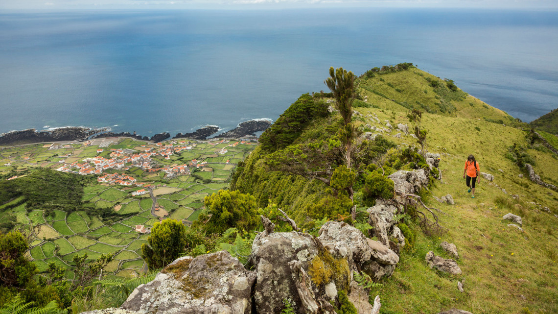 A hiker follows a grassy ridge above the west coast of Flores Island, with patchwork fields and the settlement of Faj&atilde; Grande below. The trail overlooks volcanic cliffs and the Atlantic Ocean.
