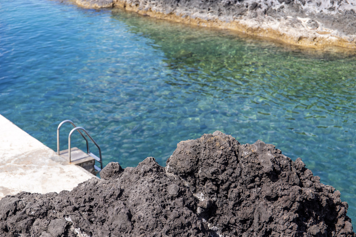 Clear seawater fills Po&ccedil;a das Salemas in Santa Cruz das Flores, with a metal ladder providing access from the volcanic shoreline. Dark basalt rocks frame the sheltered natural swimming pool.