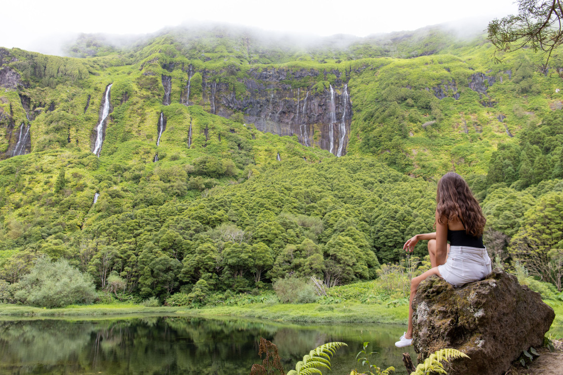 A visitor sits beside the lagoon at Po&ccedil;o da Ribeira do Ferreiro near Faj&atilde;zinha on Flores Island, Azores, Portugal. Multiple waterfalls descend from mist-covered volcanic cliffs into a densely forested valley reflected in the calm water.