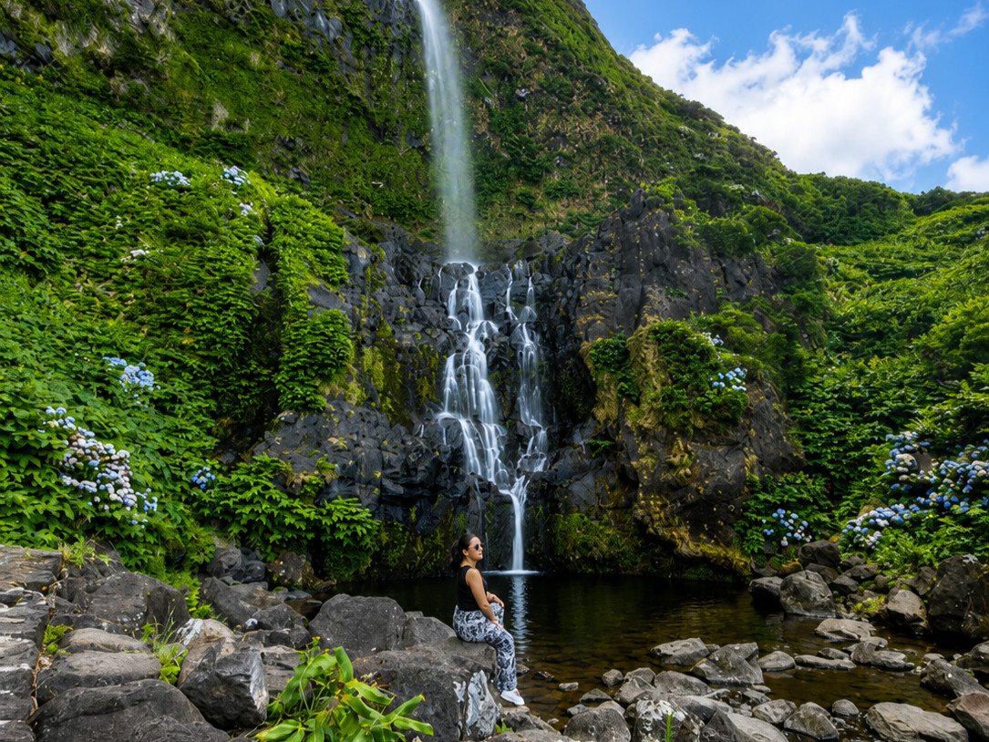 A visitor sits beside the natural pool at Cascata do Po&ccedil;o do Bacalhau in Faj&atilde; Grande on Flores Island, Azores, Portugal. The waterfall flows over dark volcanic rock surrounded by dense vegetation and blue hydrangeas.