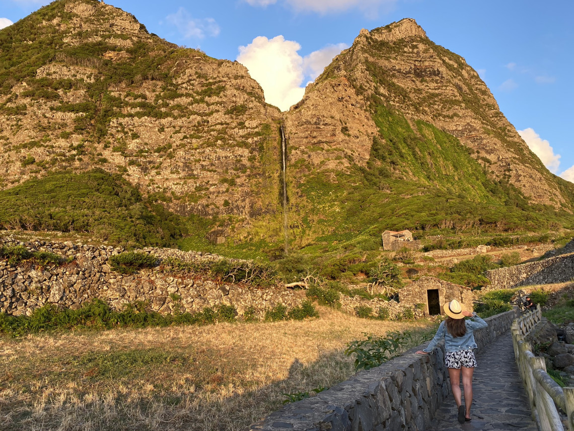 A visitor walks along the stone pathway leading to Cascata do Po&ccedil;o do Bacalhau in Faj&atilde; Grande on Flores Island, Azores, Portugal. The narrow waterfall descends between steep volcanic peaks above terraced fields and traditional stone walls.