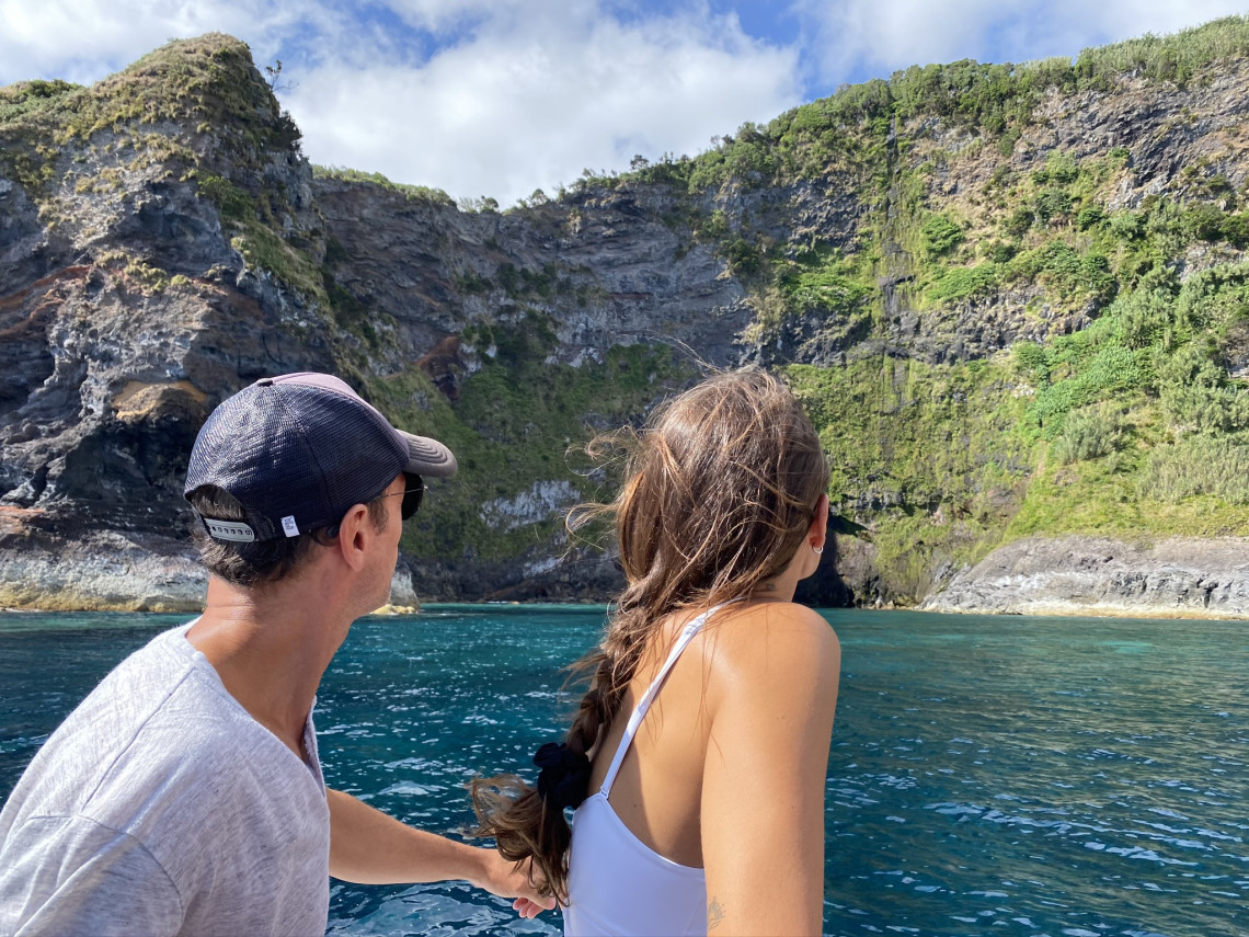 Two visitors view the steep volcanic cliffs and coastal caves of Flores Island in the Azores, Portugal, from a boat on the Atlantic Ocean. The excursion follows the island&rsquo;s rugged western coastline, where several caves and rock formations are accessible mainly by sea.