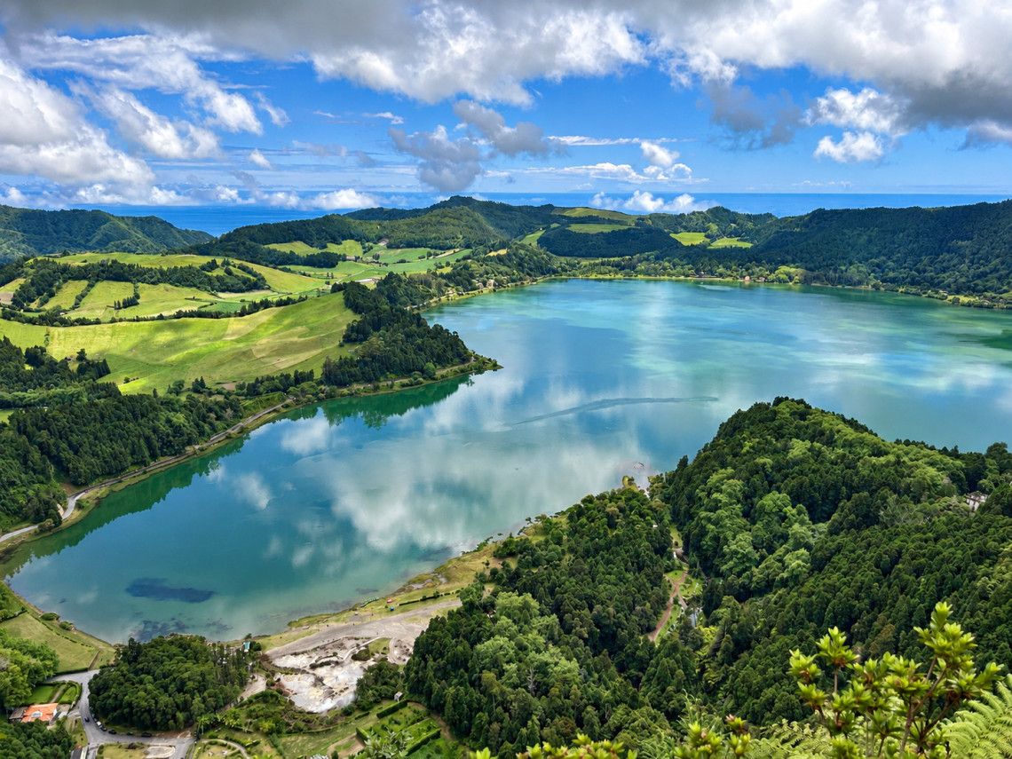 Elevated view over Lagoa das Furnas, with the volcanic lake reflecting the sky and geothermal activity visible along the shoreline beneath the surrounding green hills.