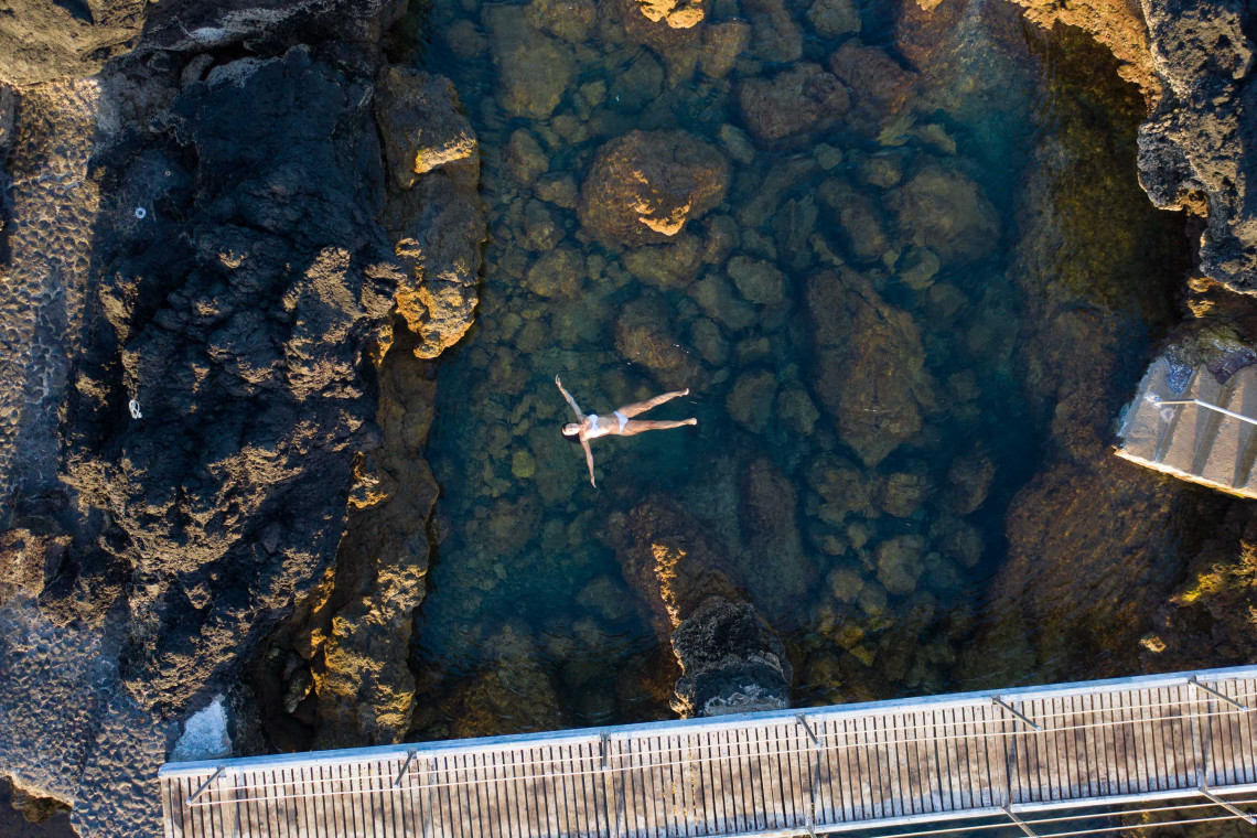 Aerial view of a swimmer in the clear seawater at Biscoitos Natural Pools on Terceira Island, surrounded by dark volcanic rocks and coastal walkways.
