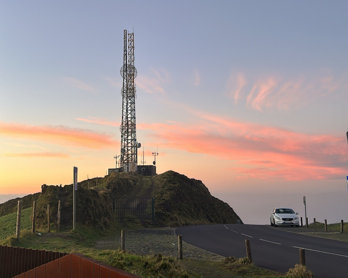 A white car navigating a winding hilltop road above Lagoa do Fogo on S&atilde;o Miguel at sunset, with a telecommunications mast on the rocky summit and a pink and orange sky over the Atlantic landscape below