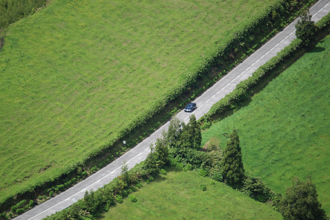 Aerial view of a single dark car driving on a narrow road flanked by hedgerows, cutting diagonally through bright green Azorean pasture land