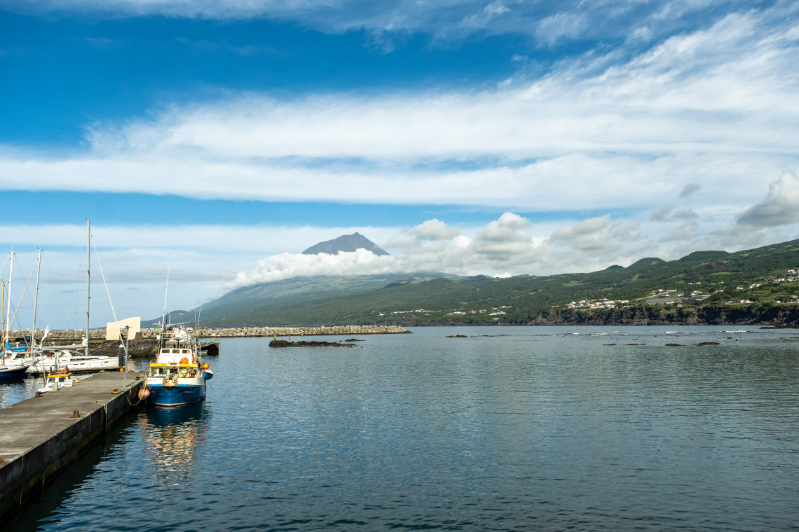Madalena harbour on Pico Island, with fishing boats and sailing vessels moored at the pier, Mount Pico's volcanic cone partially hidden by clouds rising steeply behind, and the green coastal slopes of the island stretching to the right