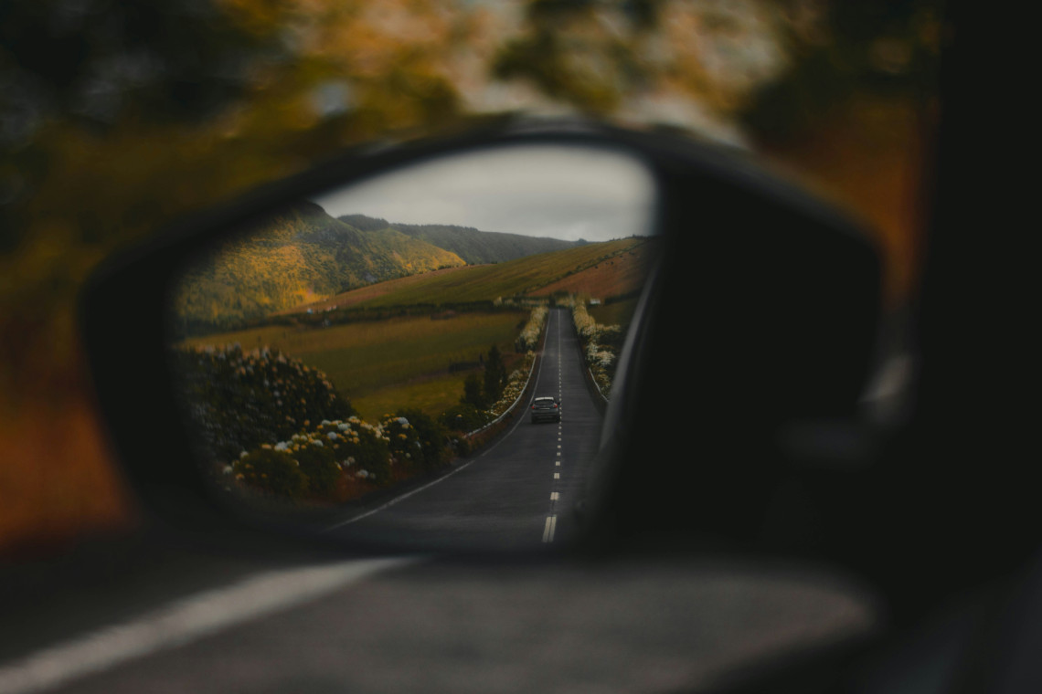 A car's wing mirror in sharp focus reflecting a straight open road receding through green and golden Azorean hillside landscape under an overcast sky, with another car visible in the distance