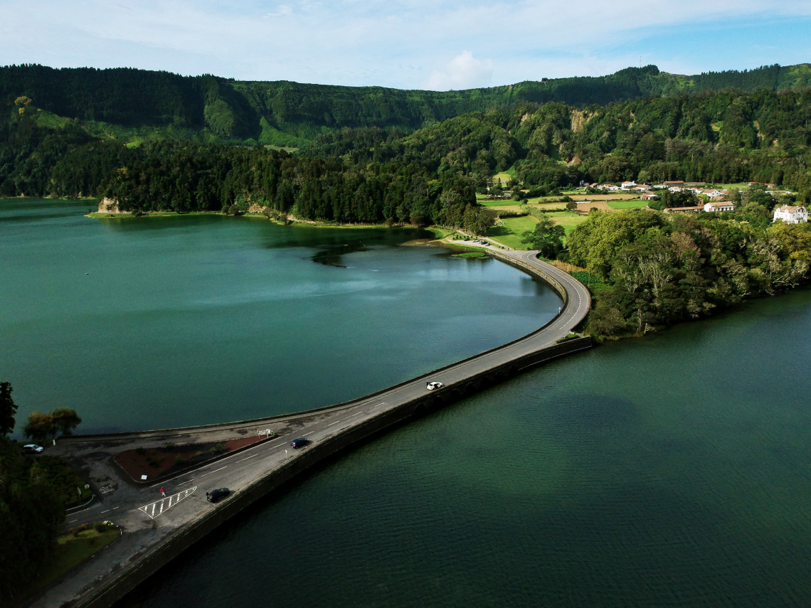 Aerial view of the narrow causeway road separating the two crater lakes of Sete Cidades on S&atilde;o Miguel, with the green Lagoa Verde on the left, cars on the road, dense forested crater walls rising behind, and the village and farmland visible on the far shore