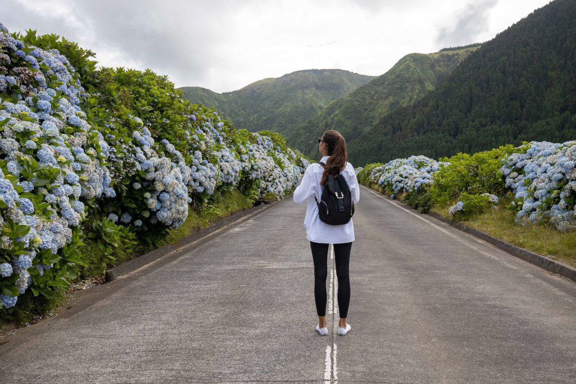 Viajera caminando por una carretera rodeada de hortensias azules y monta&ntilde;as verdes en las Azores, un entorno natural ideal para recorrer la isla con libertad