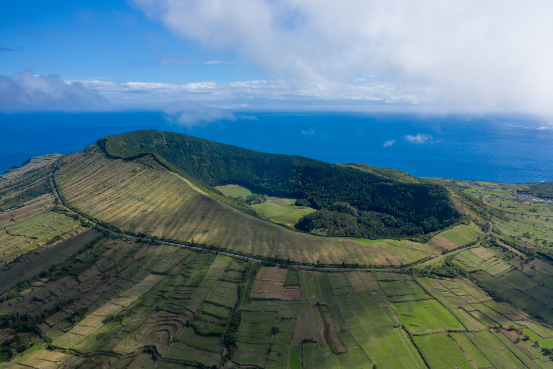 Aerial view of Caldeira da Graciosa, showing its forested volcanic crater bordered by agricultural fields and the blue Atlantic Ocean.