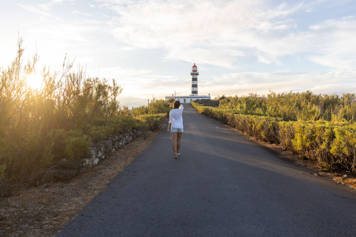 Traveler walking toward the black-and-white Ponta da Barca Lighthouse on Graciosa Island, surrounded by low coastal vegetation in warm evening light.