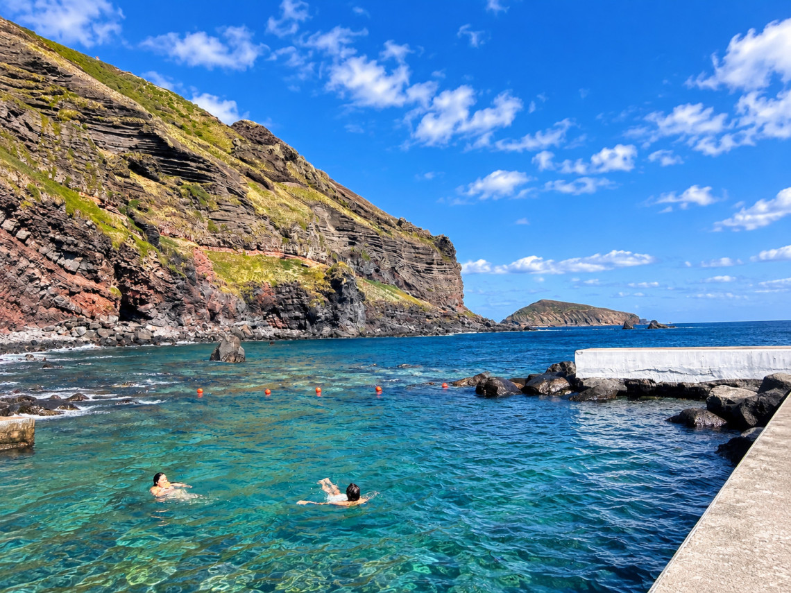Swimmers enjoying the clear seawater at the Carapacho Natural Pools on Graciosa Island, surrounded by layered volcanic cliffs and dark basalt formations.