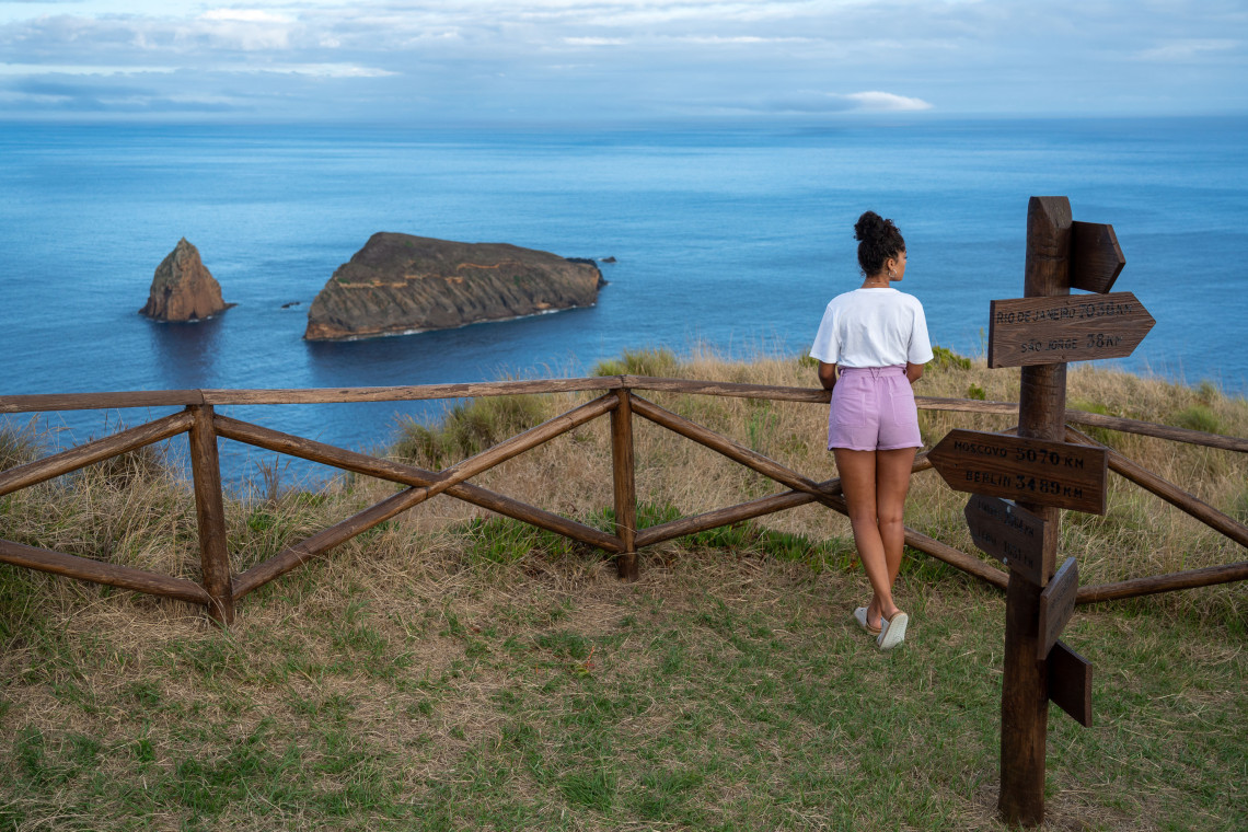 Traveler overlooking the Atlantic from Ponta da Restinga Viewpoint on Graciosa Island, with the rocky Ilh&eacute;us do Carapacho visible offshore beside a wooden signpost.