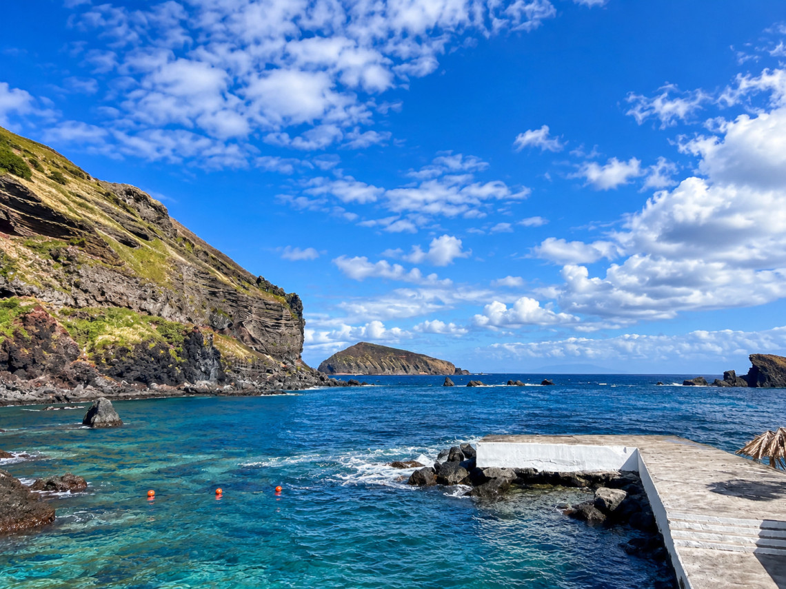 Clear seawater at the Carapacho Natural Pools on Graciosa Island, framed by dark volcanic rock, steep layered cliffs and offshore formations beneath a bright blue sky.
