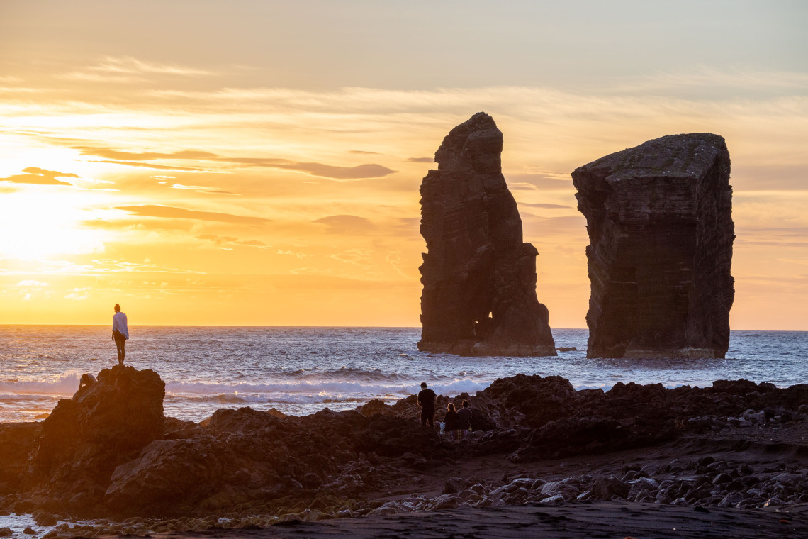 Sunset over the volcanic Mosteiros Islets off the western coast of S&atilde;o Miguel Island, with visitors standing among dark coastal rocks beside the Atlantic Ocean.