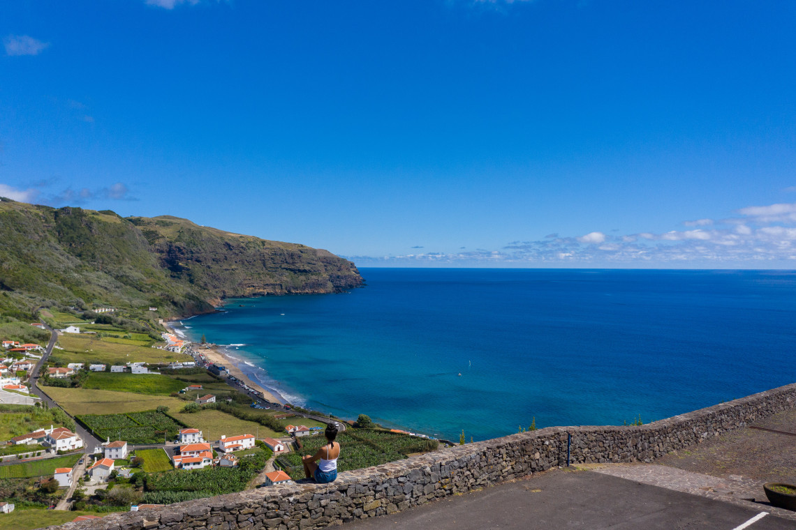 Elevated view over Praia Formosa on Santa Maria Island, showing the sheltered Atlantic bay, coastal village, green fields and steep volcanic cliffs.