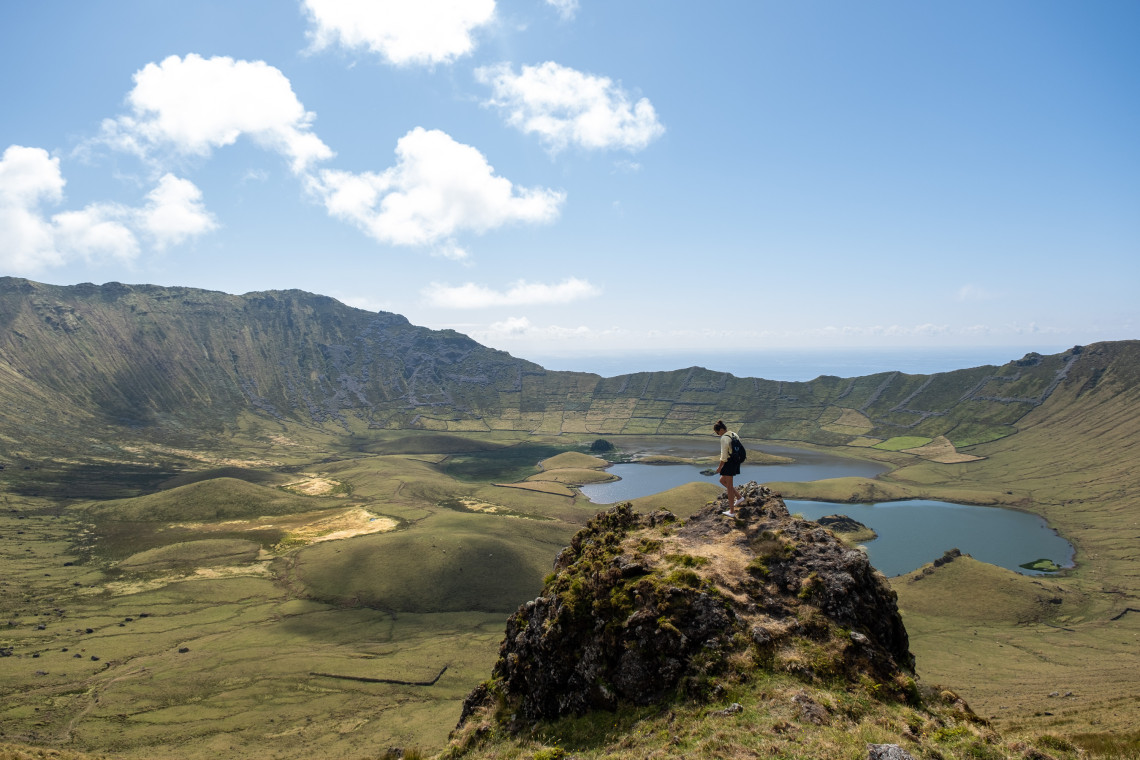 A hiker stands on a rocky outcrop overlooking Caldeir&atilde;o Crater on Corvo Island in the Azores, Portugal. Small lakes and volcanic cones spread across the green floor of the island&rsquo;s central caldera.