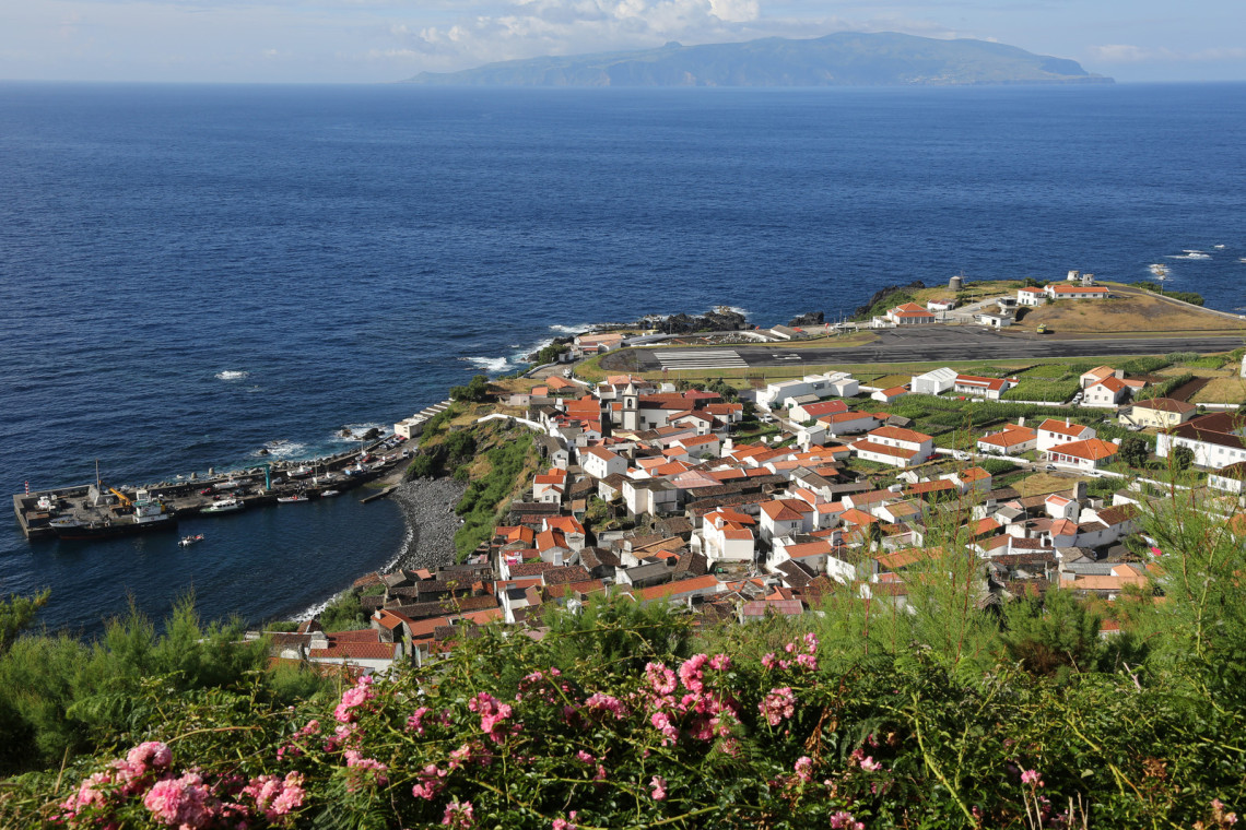 Elevated view of Vila do Corvo and its harbour on Corvo Island in the Azores, Portugal. White houses with red-tiled roofs cluster beside the Atlantic, with Flores Island visible across the water.