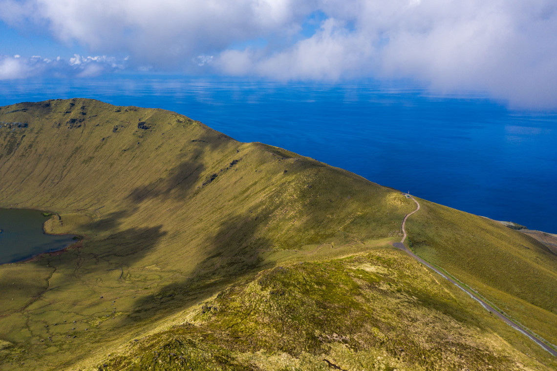 View along the grassy rim of Caldeir&atilde;o Crater on Corvo Island in the Azores, Portugal, with part of the crater lake visible below. A narrow road crosses the volcanic highlands with the Atlantic Ocean extending beyond the island.