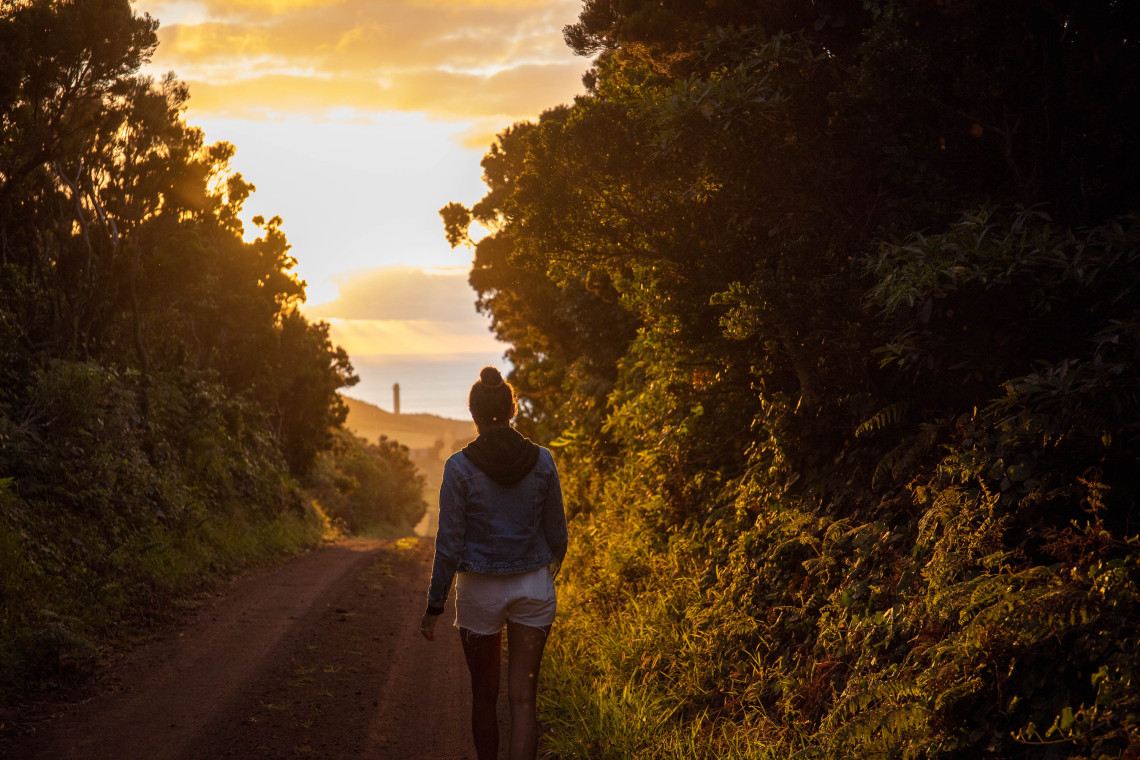 Traveler walking along a rural track at Ponta dos Rosais on S&atilde;o Jorge Island, with Rosais Lighthouse visible ahead beneath the warm evening sky.