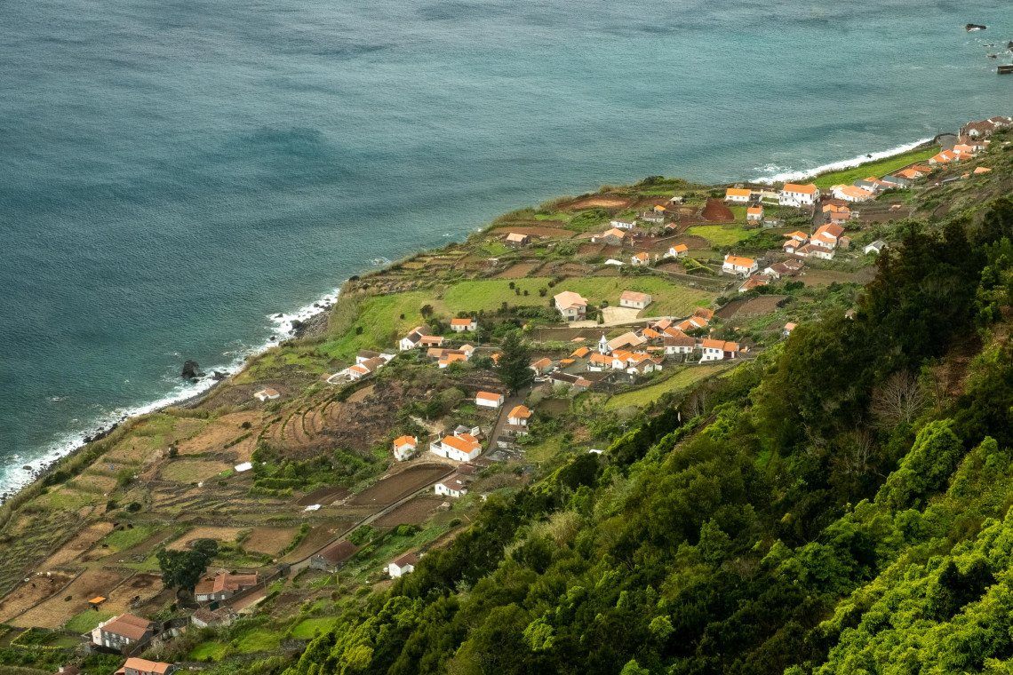 Elevated view of a traditional coastal faj&atilde; on S&atilde;o Jorge Island, with terraced agricultural plots, white houses with red roofs and the Atlantic Ocean along the shoreline.