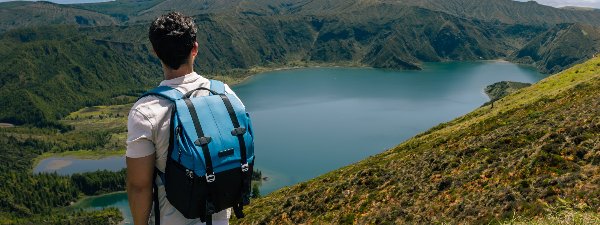 Hiker overlooking Lagoa do Fogo on São Miguel Island, with the blue crater lake and steep green volcanic slopes extending across the landscape.