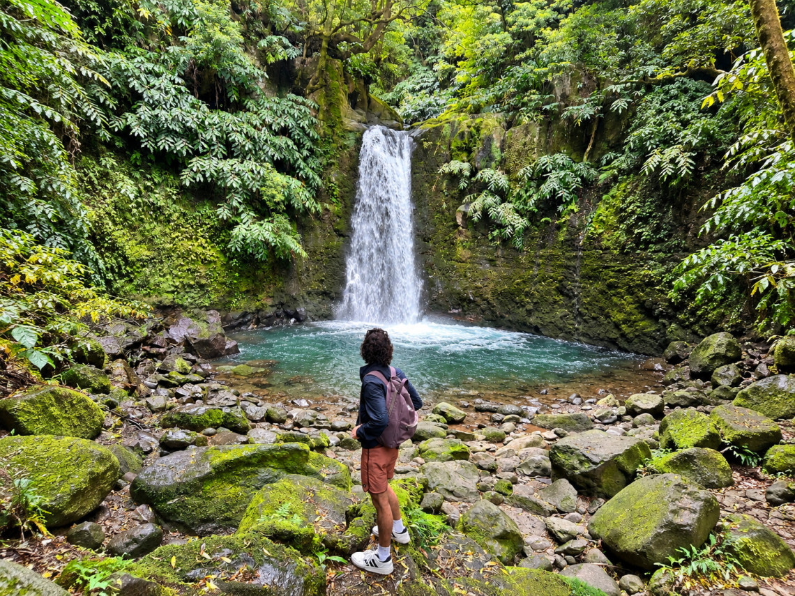 Hiker standing in front of Salto do Prego waterfall in Faial da Terra, surrounded by moss-covered rocks and dense forest vegetation on S&atilde;o Miguel Island.