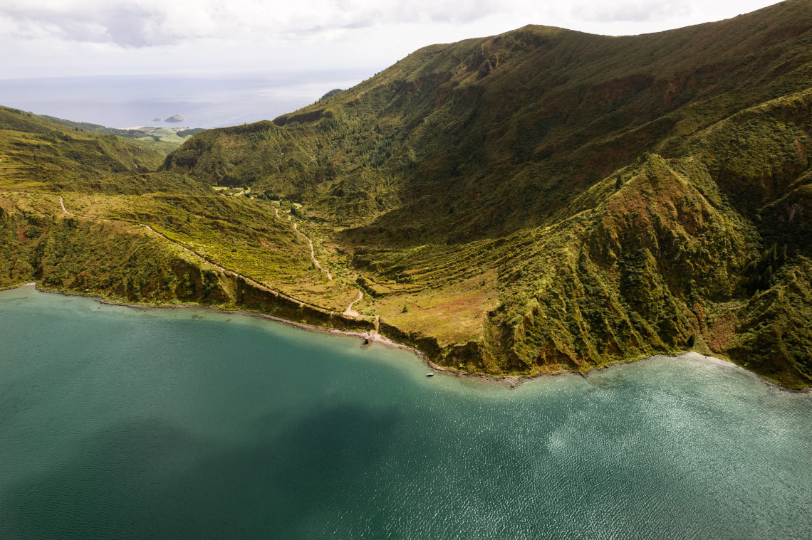 Aerial view of a hiking trail along the shores of Lagoa do Fogo, surrounded by steep volcanic ridges and the Atlantic landscape of S&atilde;o Miguel Island.