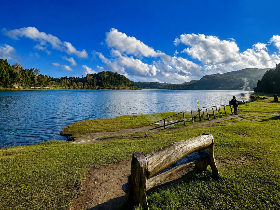 Peaceful lakeside view at Lagoa das Furnas on S&atilde;o Miguel Island, with a wooden bench, grassy shoreline and forested hills beneath a bright blue sky.