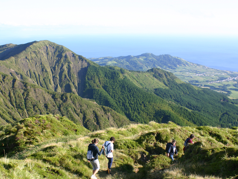 Hikers descending a trail near Pico da Vara on S&atilde;o Miguel Island, surrounded by steep volcanic mountains, green forest and views toward the Atlantic coast.