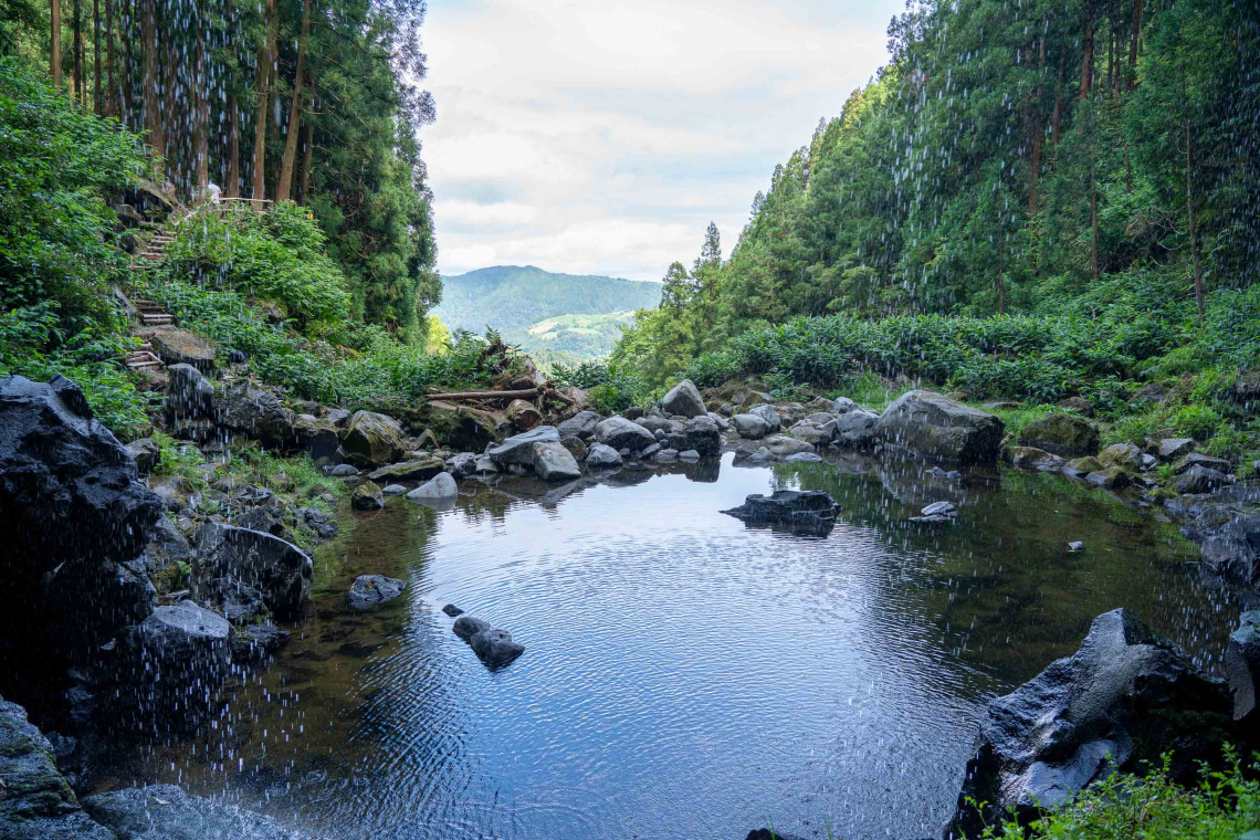 Visitors standing beside a waterfall in Gren&aacute; Natural Park in Furnas, surrounded by volcanic rock, forest vegetation and a natural stream.
