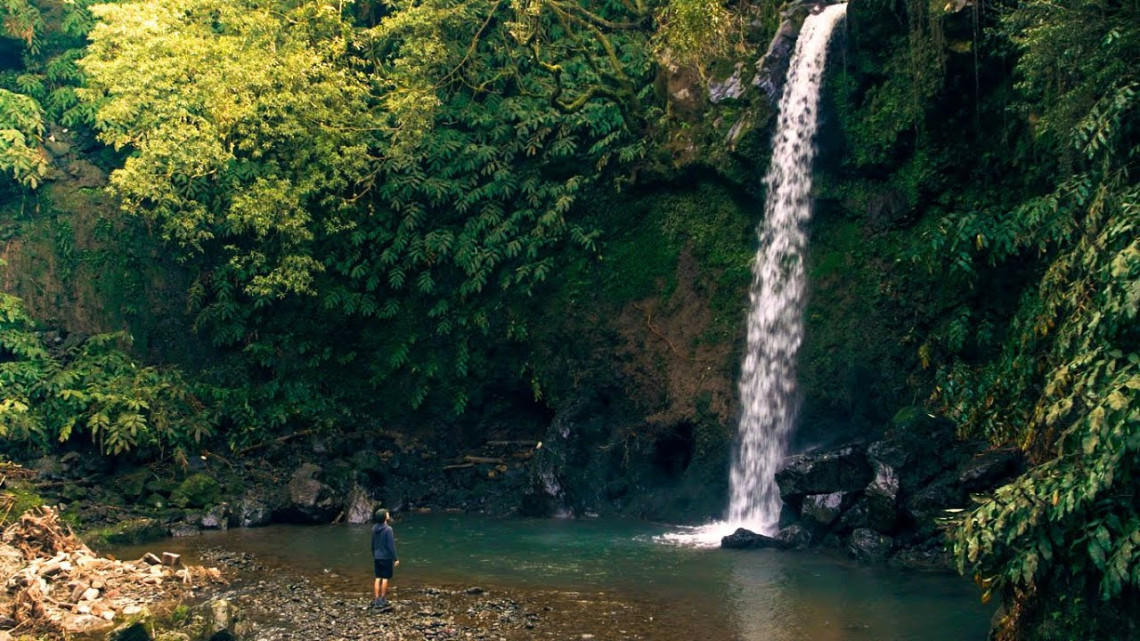 Visitor swimming in a clear natural pool at Moinhos do F&eacute;lix on S&atilde;o Miguel Island, beneath a small waterfall surrounded by volcanic rock and lush vegetation.