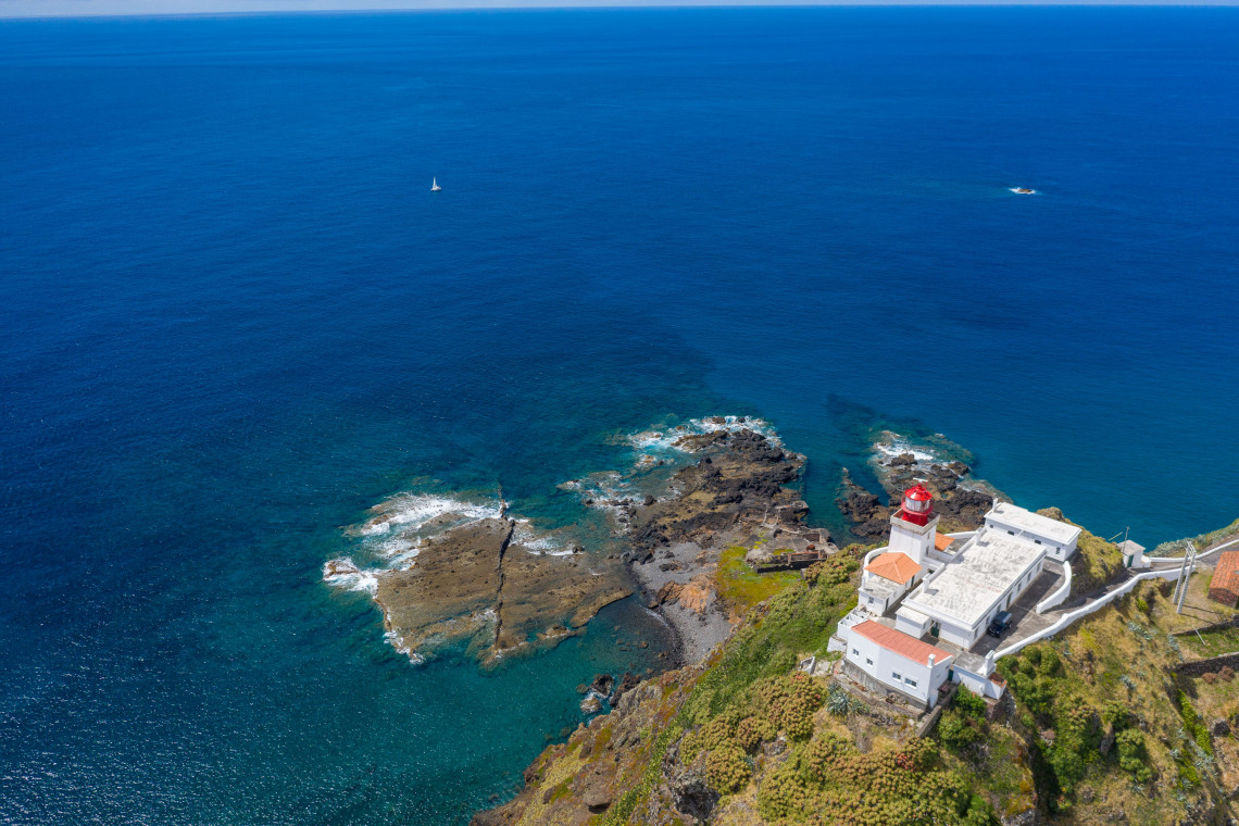 Aerial view of Gon&ccedil;alo Velho Lighthouse overlooking the Atlantic Ocean at Ponta do Castelo on Santa Maria Island, with white buildings, a red lighthouse tower and dark volcanic rocks extending into clear blue waters.
