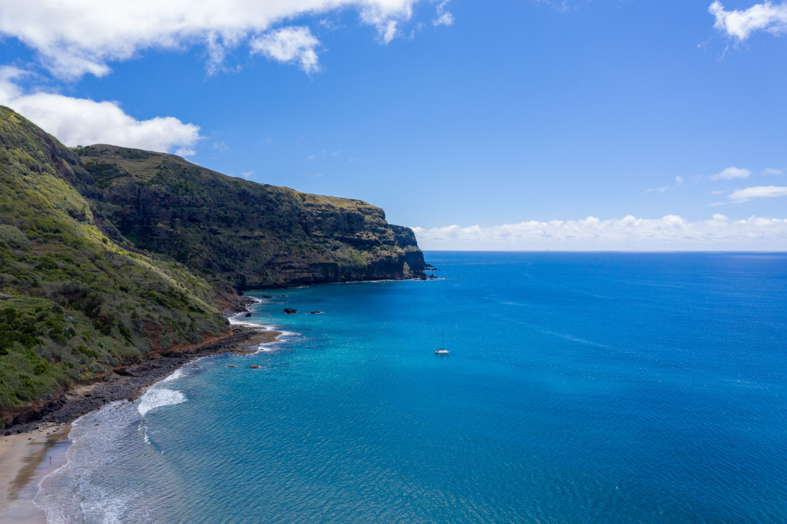 View of a sandy beach and turquoise Atlantic waters at Praia Formosa on Santa Maria Island, with green volcanic cliffs along the coast and a small sailboat anchored offshore.