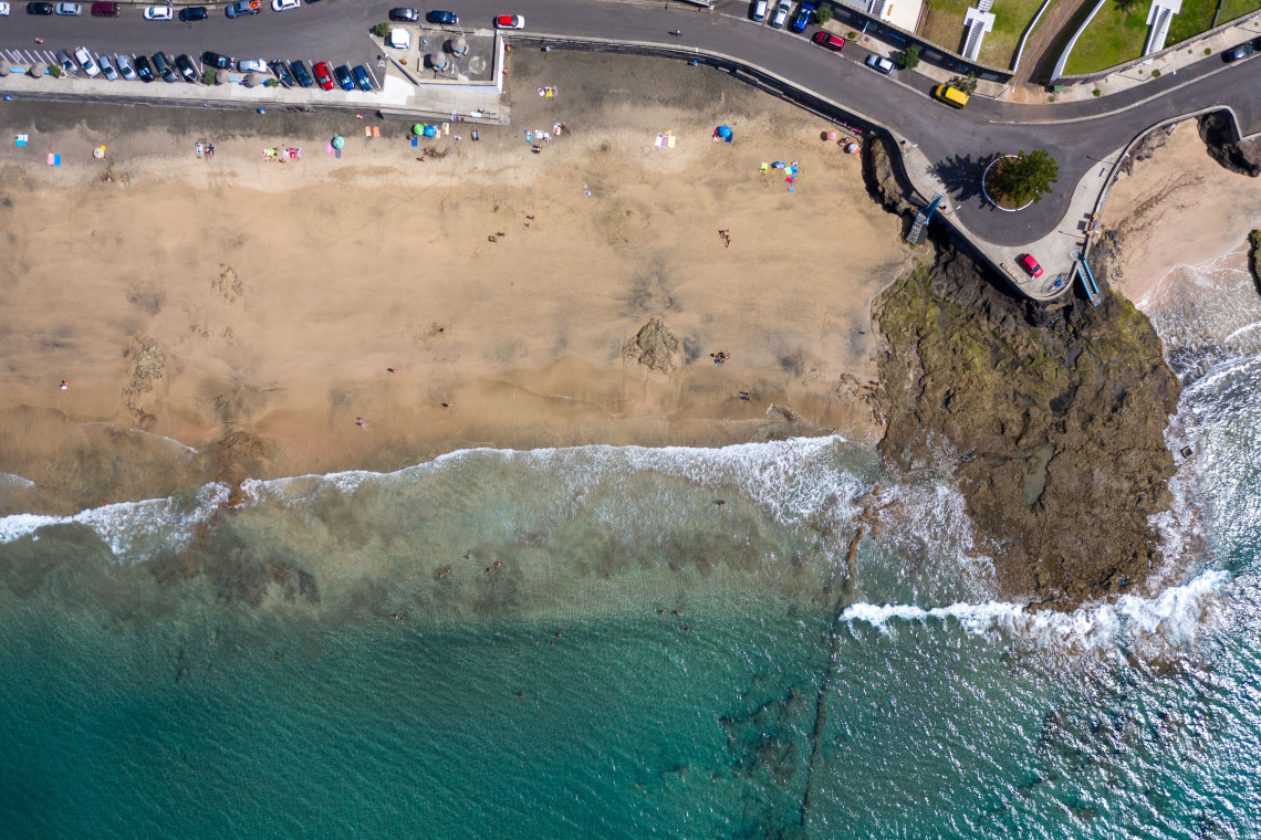 Overhead aerial view of Praia Formosa on Santa Maria Island, showing its sandy beach, visitors along the shore, a coastal road and clear Atlantic waters washing over submerged rocks.