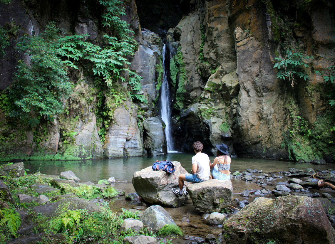 Visitors sit on rocks facing Salto do Cabrito waterfall near Ribeira Grande, surrounded by steep volcanic walls and dense vegetation.