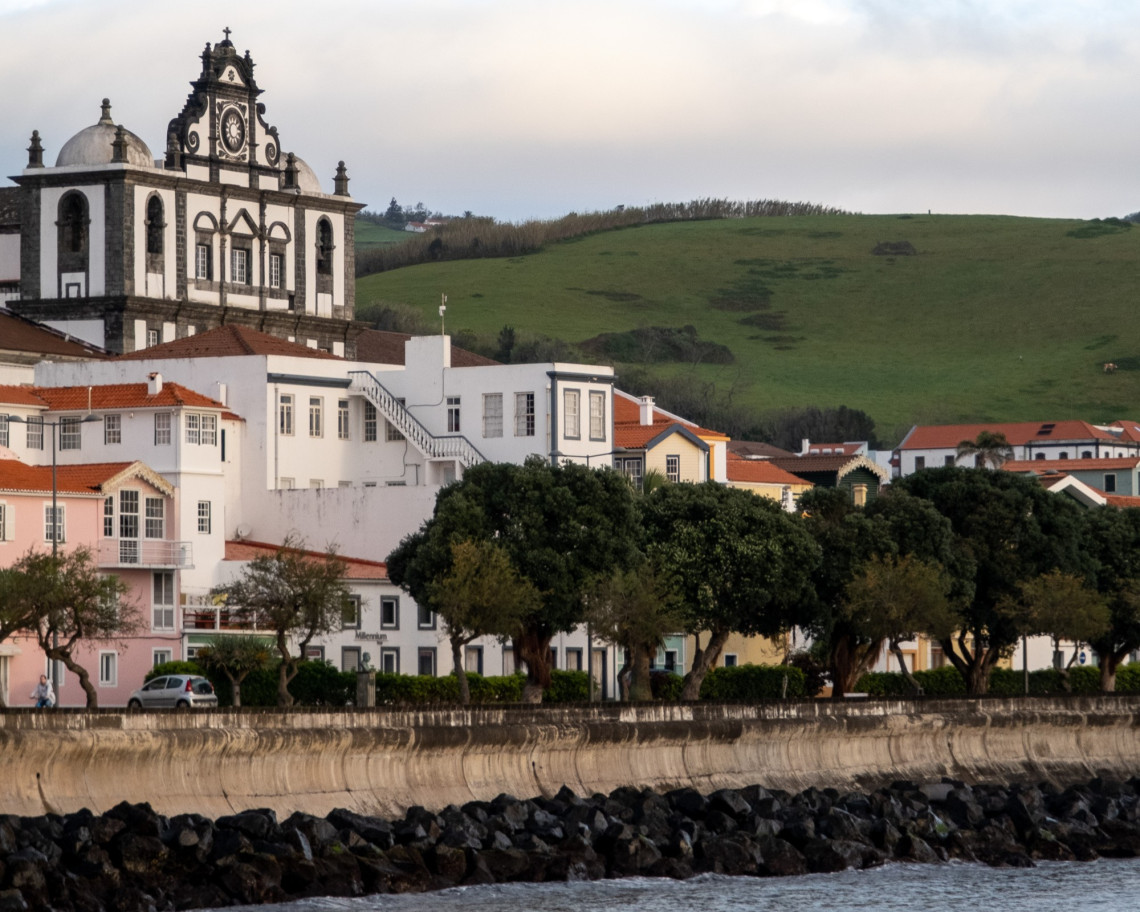 Nossa Senhora das Ang&uacute;stias Church rising above traditional buildings along the waterfront of Horta on Faial Island, with green hills in the background.