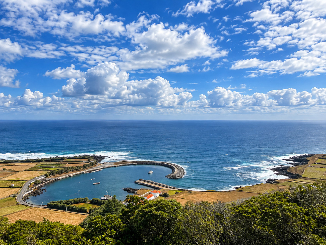 Elevated view of Porto da Barra on Graciosa Island, showing its curved protective breakwater, small harbor and surrounding agricultural landscape beside the Atlantic Ocean.