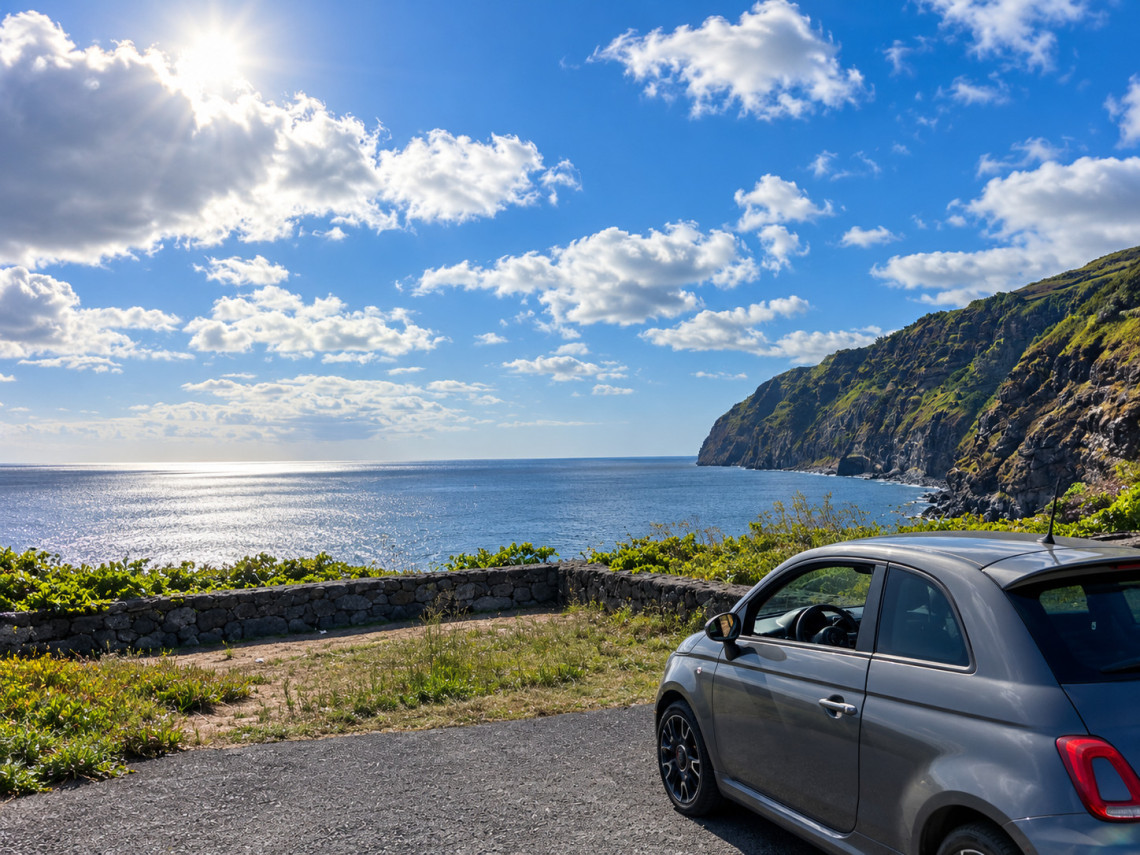 Coastal road on Graciosa Island overlooking the Atlantic Ocean, with a car beside dark volcanic stone walls and steep green cliffs extending along the shoreline.