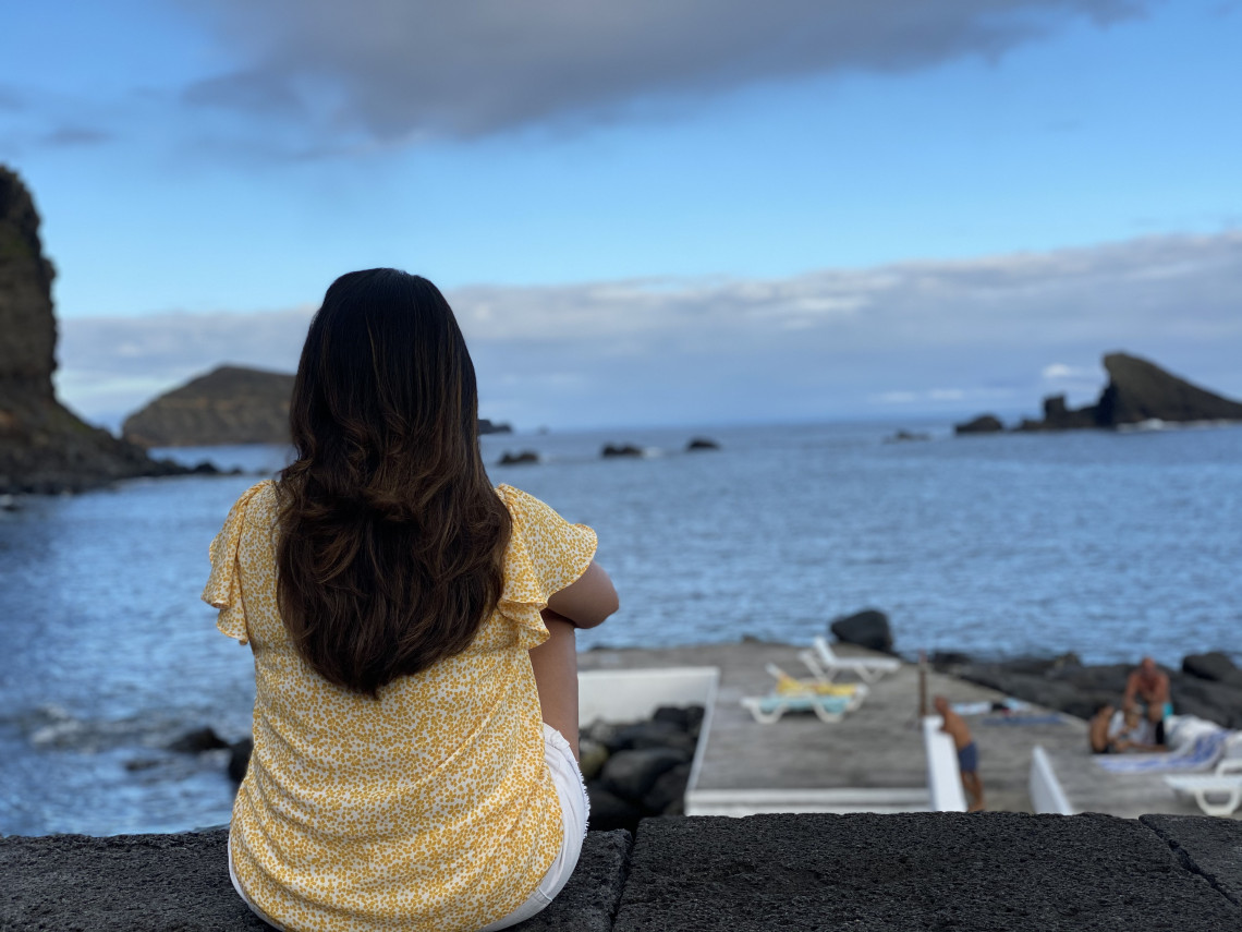 Traveler overlooking the Atlantic from the Carapacho Natural Pools on Graciosa Island, with dark volcanic rocks and offshore formations surrounding the coastal swimming area.