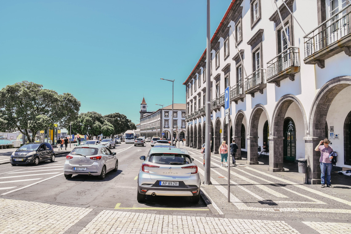 Street view in central Ponta Delgada showing traditional white buildings with dark volcanic stone details, patterned pavement and everyday city traffic.
