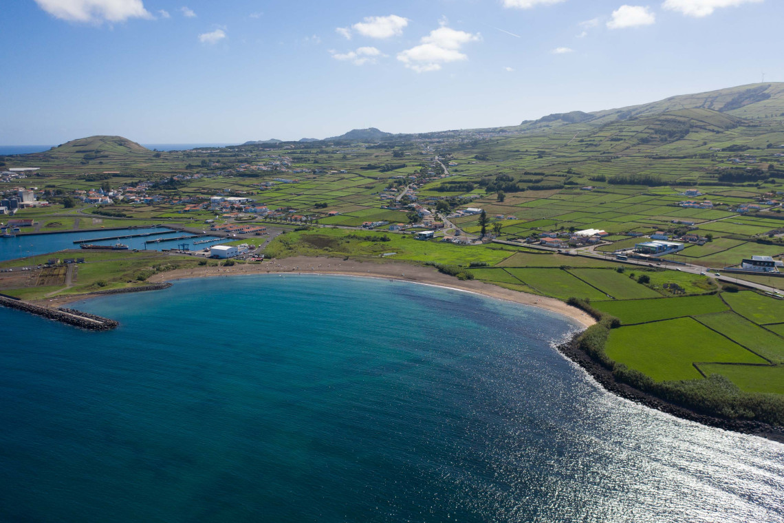 Aerial view of the coastline around Praia da Vit&oacute;ria on Terceira Island, with a curved beach, blue Atlantic waters and green agricultural fields extending toward the surrounding hills.