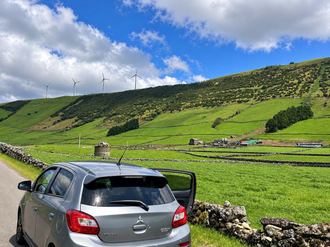 Green pastures divided by volcanic stone walls near Serra do Cume on Terceira Island, with wind turbines lining the surrounding hills. A rural road passes through the agricultural landscape beneath a blue and cloudy Azorean sky.