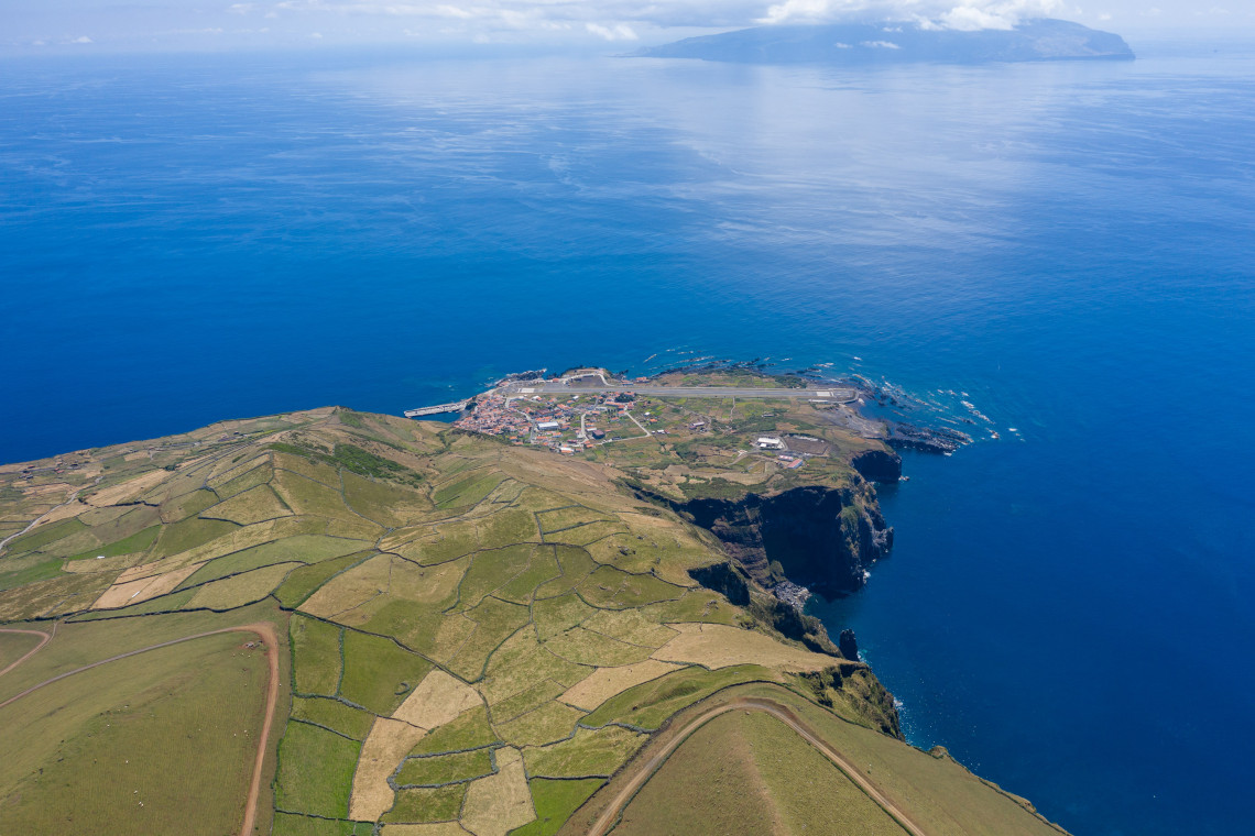 Elevated view over patchwork farmland leading toward Vila do Corvo on Corvo Island, with the coastal settlement, volcanic cliffs, and Atlantic Ocean below.