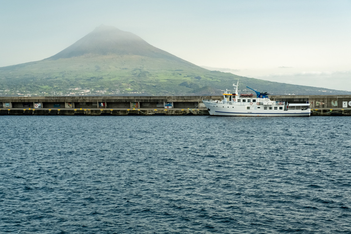 Ferry docked at Madalena Harbor on Pico Island in the Azores, with Mount Pico rising behind the waterfront and harbor wall.