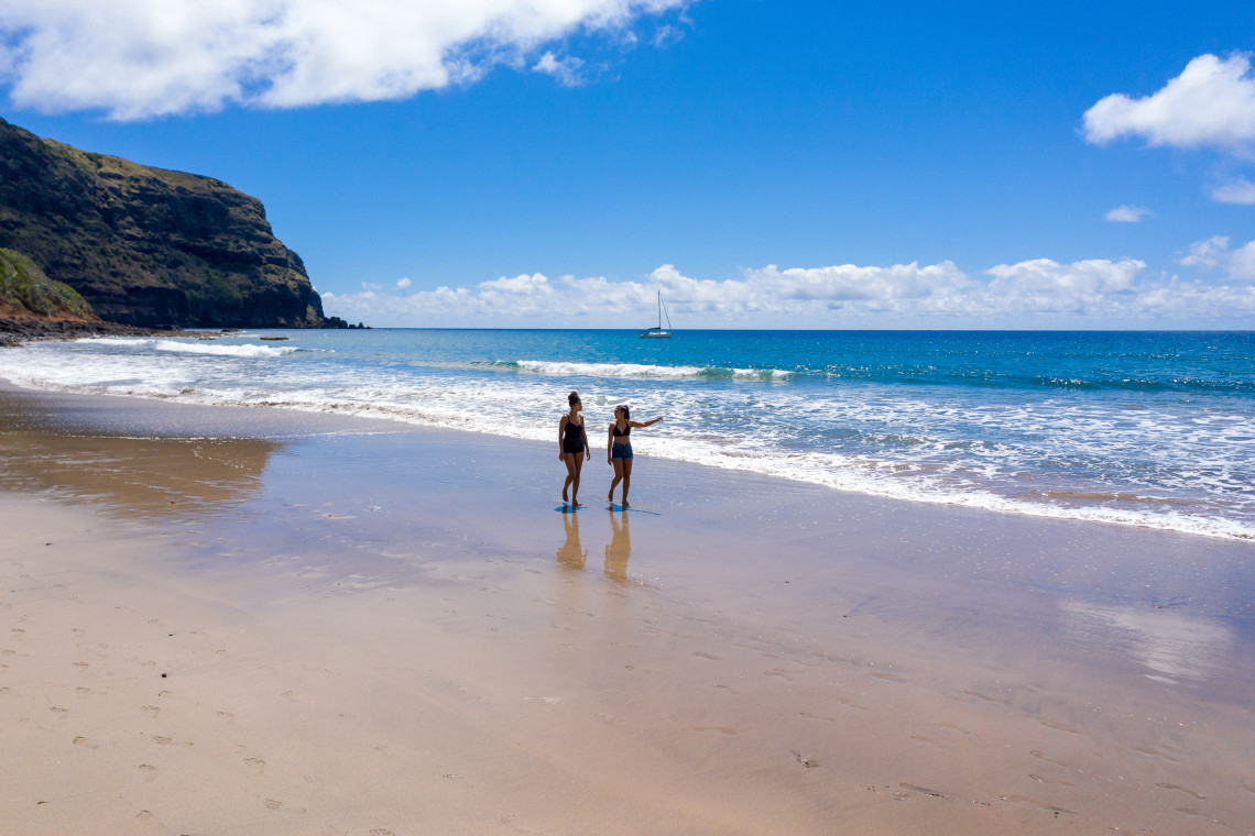 Two visitors walk along the sandy shoreline of Praia Formosa on Santa Maria Island, with Atlantic waves, a green coastal cliff and a sailboat offshore.