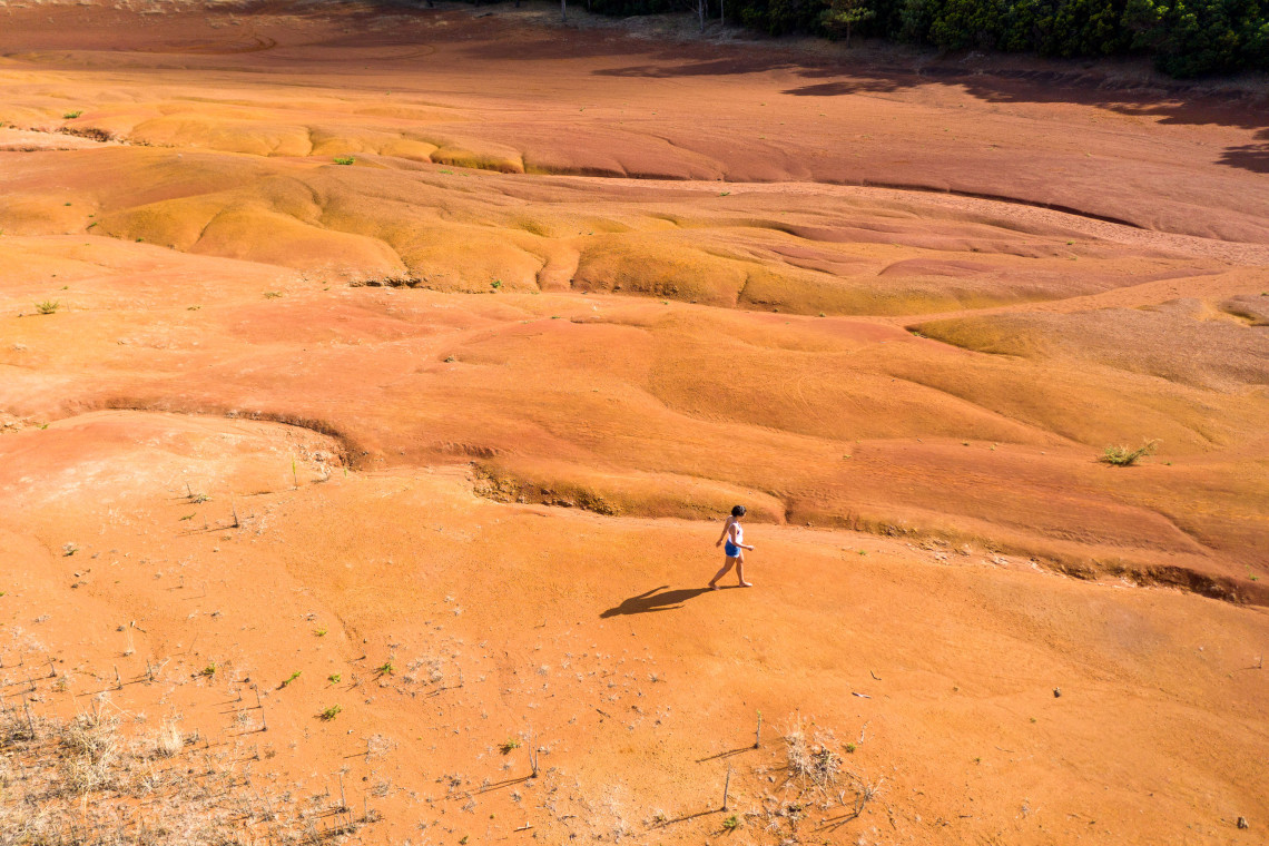 A visitor walks across the reddish clay terrain of Barreiro da Faneca on Santa Maria Island, surrounded by eroded hills and sparse vegetation.